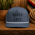 Gray Rodeo Loco western rope hat with black embroidery displayed on rustic wooden table with western decor background.