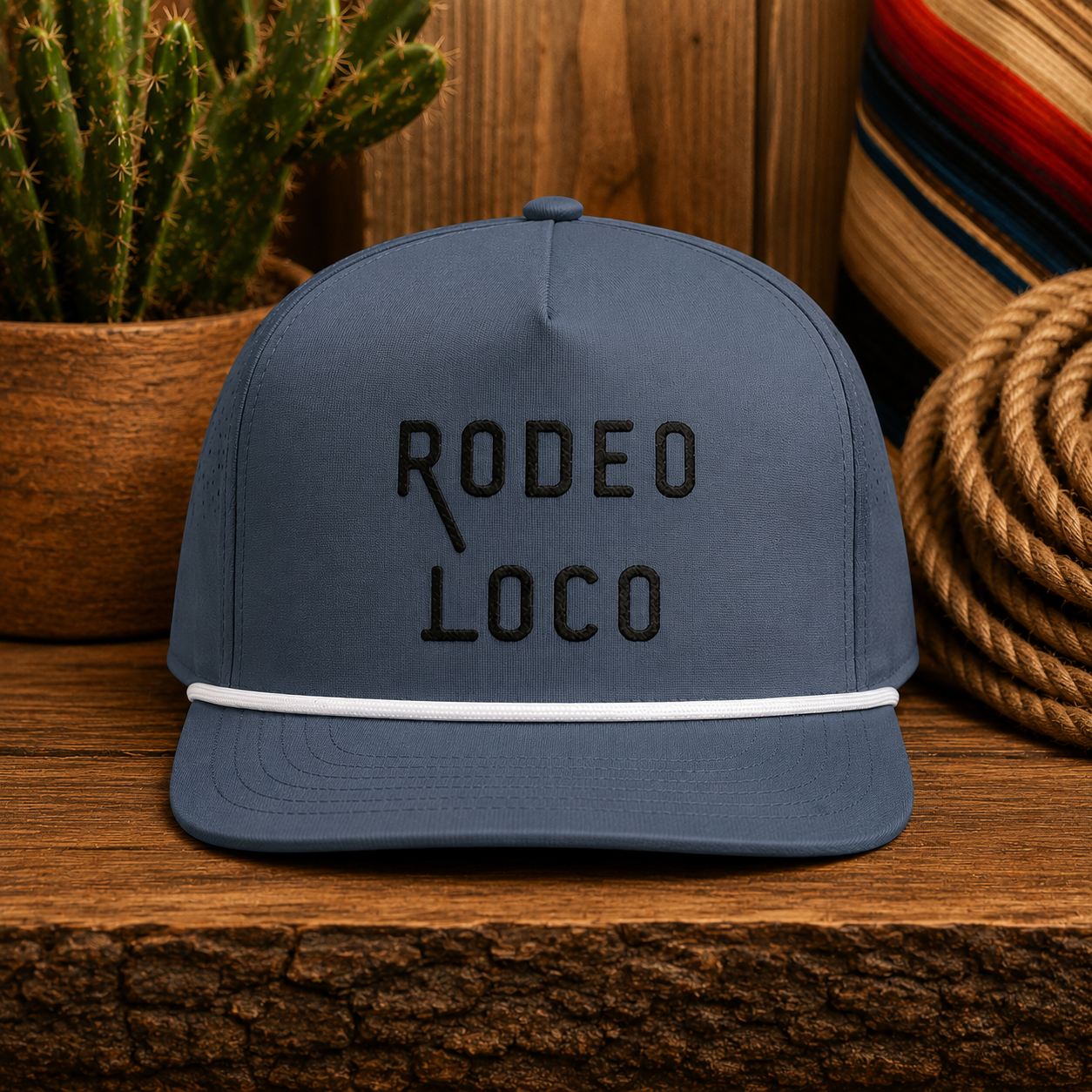 Gray Rodeo Loco western rope hat with black embroidery displayed on rustic wooden table with western decor background.
