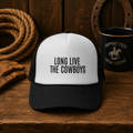 Long Live The Cowboys western trucker hat with black and white design, displayed on a rustic wooden table with western ranch decor.
